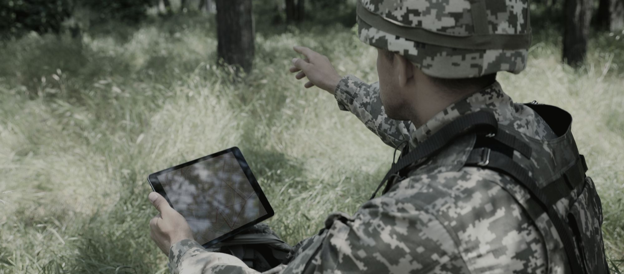 Militar em área de mata consultando um tablet e indicando a direção ao redor.