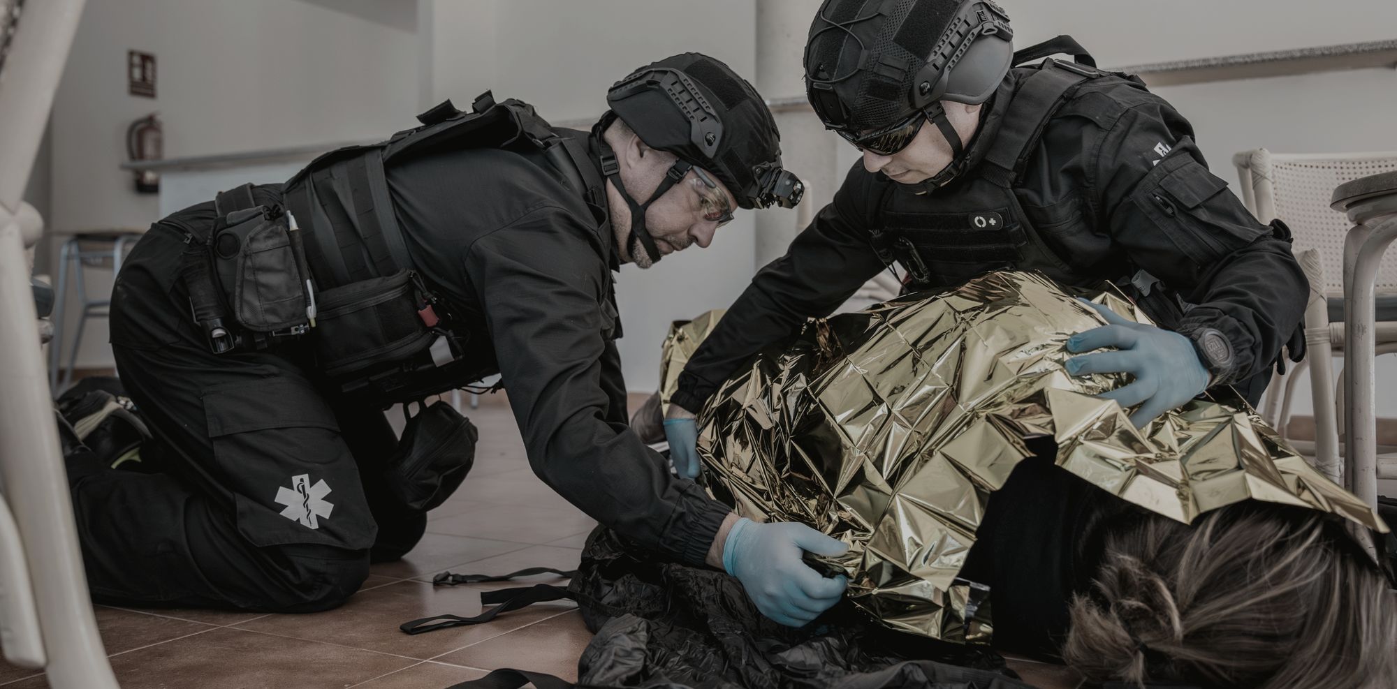 Dois socorristas de equipe tática atendendo juntos uma vítima simulada coberta por manta térmica.