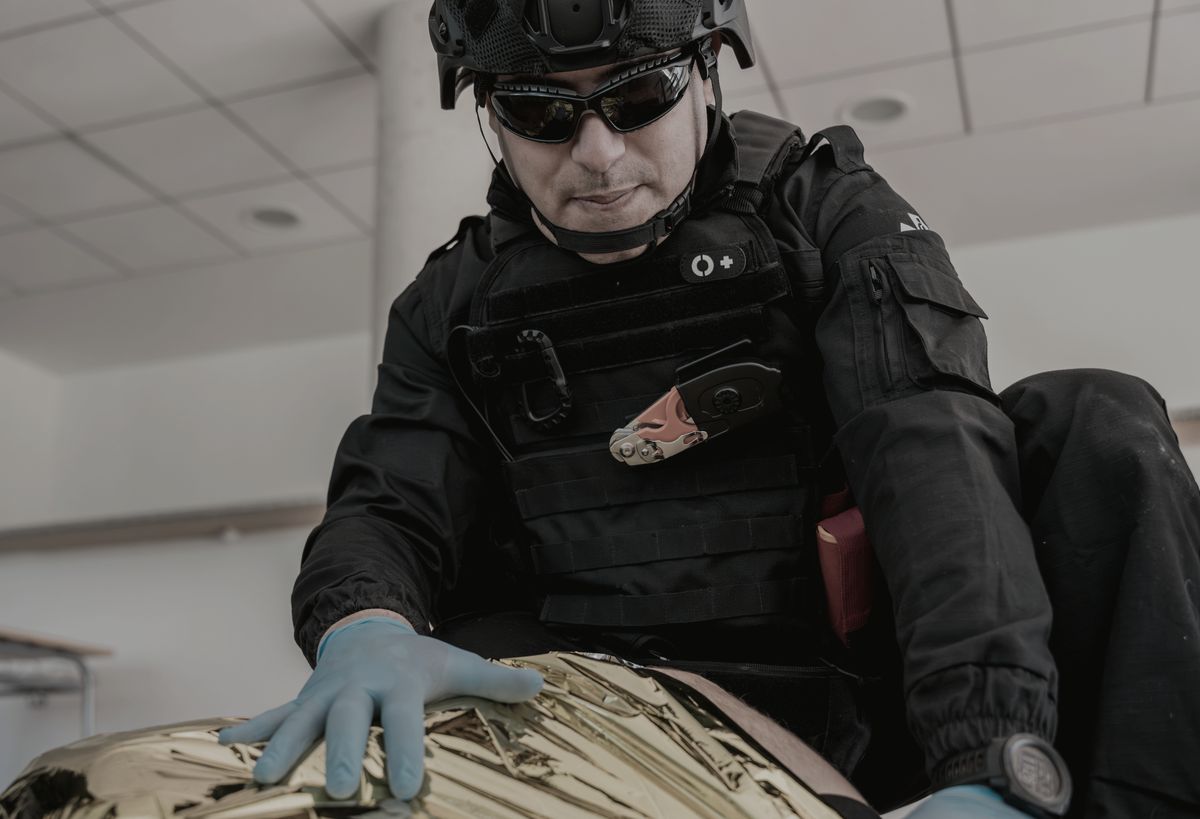 Instrutor com capacete e colete atendendo uma vítima simulada coberta por manta térmica em sala de treinamento.